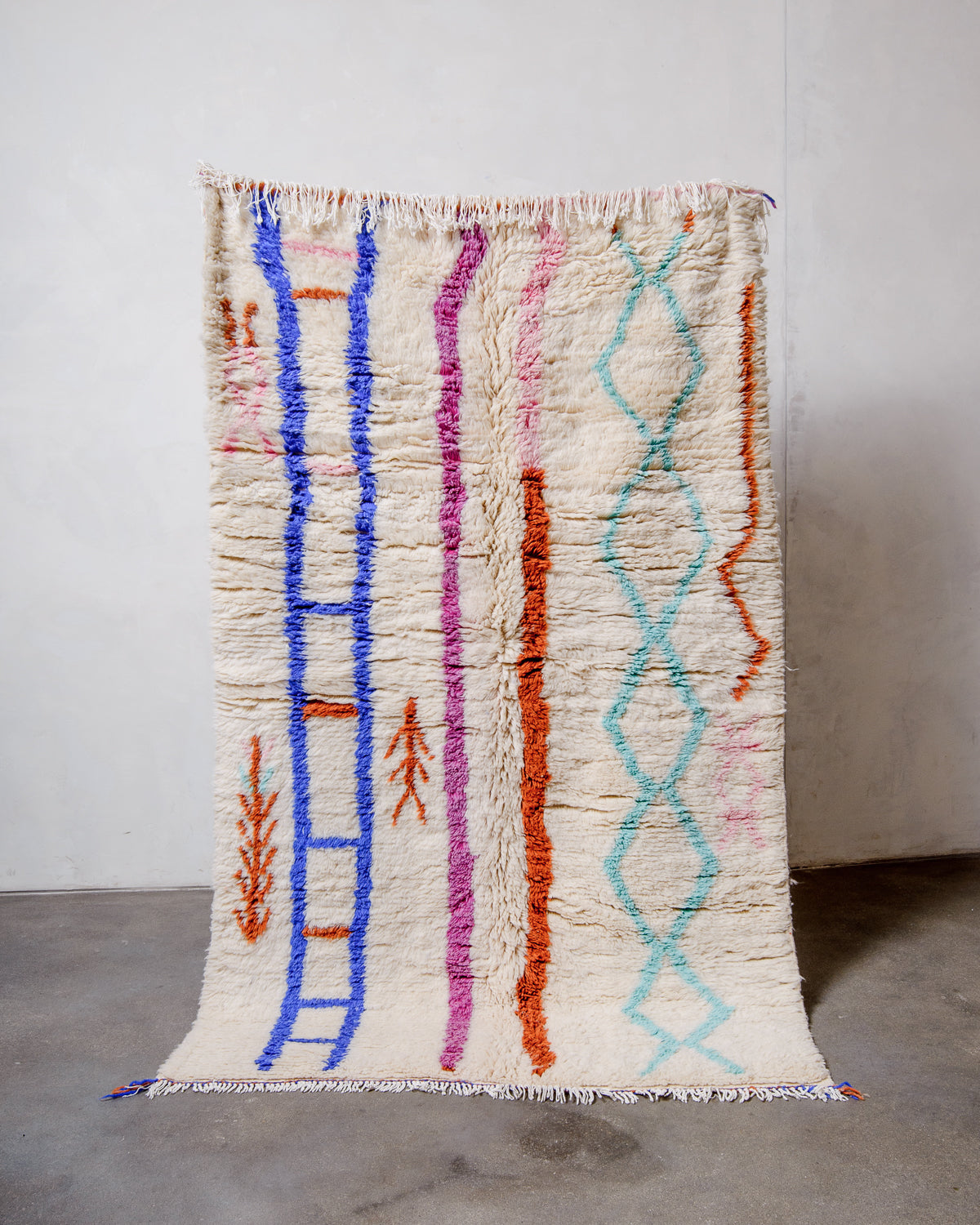 Berberlin Azilal rug made of wool with cotton tassels, held up against a plain white wall and cement floor.